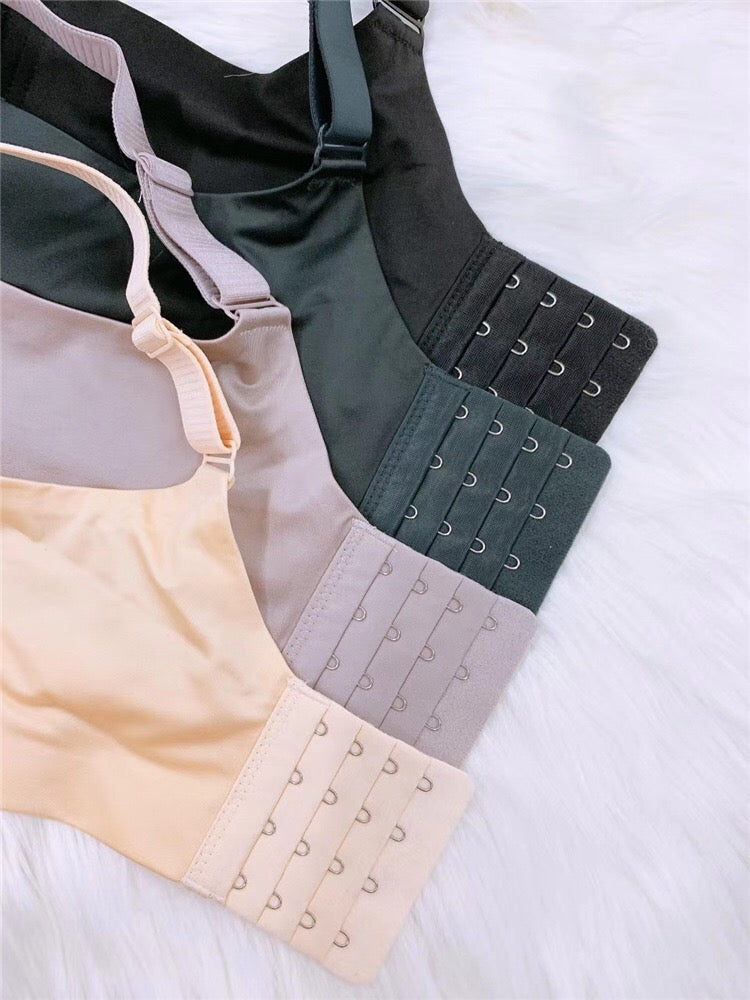 A close-up of four bras in different colors (black, green, beige, and light gray) laid out on a soft white fabric. The bras feature wide, adjustable back straps with multiple hook-and-eye closures for added support and a customizable fit.